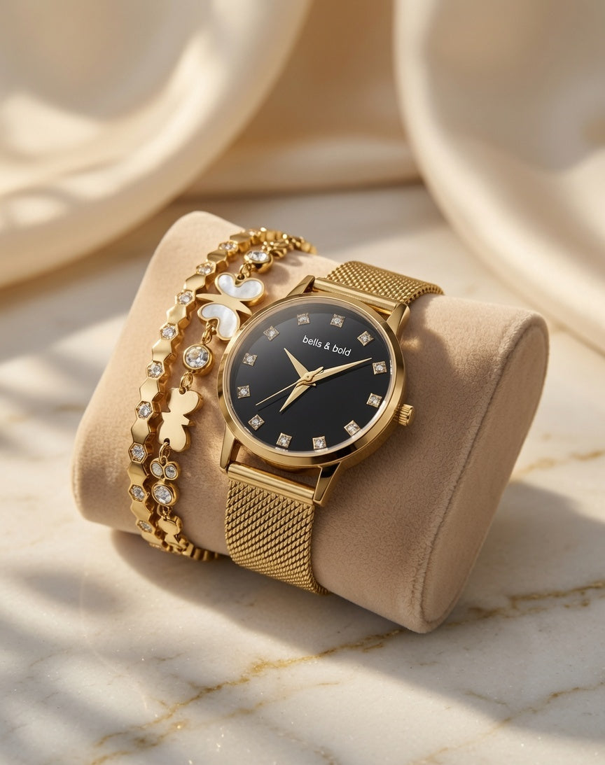 Gold watch with black face and diamond accents on a beige cushion, surrounded by jewelry on a marble surface.